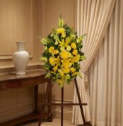 Pedestal de homenaje "El Legado de Sebastian" con un arreglo floral fúnebre de lirios y crisantemos amarillos, presentado ele