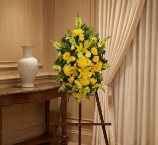 Pedestal de homenaje "El Legado de Sebastian" con un arreglo floral fúnebre de lirios y crisantemos amarillos, presentado ele