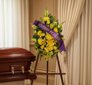 Pedestal de homenaje "El Legado de Sebastian" con flores amarillas y cinta morada "Sentido Pésame" junto a un ataúd en sala d
