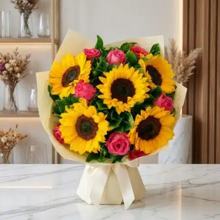 Ramillete o bouquet de girasoles y rosas rosadas, envuelto en papel crema con lazo, sobre mesa de mármol en floristería premi