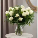 Ramo de 18 rosas blancas frescas en un jarrón de cristal, con follaje verde, sobre una mesa de mármol y fondo elegante.