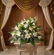 Ramo fúnebre Abdiel con lirios blancos y follaje verde, presentado elegantemente en una sala de velación con cortinas doradas