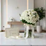 Ramo JACOBELLA de 60 rosas blancas en jarrón de cristal con cinta satinada, tarjeta de regalo y sobre sobre superficie de már