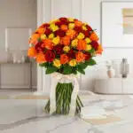 Ramo SYLVANA con 60 rosas rojas, naranjas y amarillas, atado con cinta de satén crema, sobre mesa de mármol con tarjeta de re