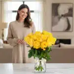 Mujer sonriente admirando un arreglo floral de 18 rosas amarillas en un jarrón de cristal con lazo, en un hogar de diseño luj