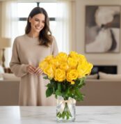 Mujer sonriente admirando un arreglo floral de 18 rosas amarillas en un jarrón de cristal con lazo, en un hogar de diseño luj
