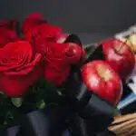 Primer plano de rosas rojas frescas con gotas de rocío, manzanas rojas brillantes y un lazo negro de satén en una cesta de re